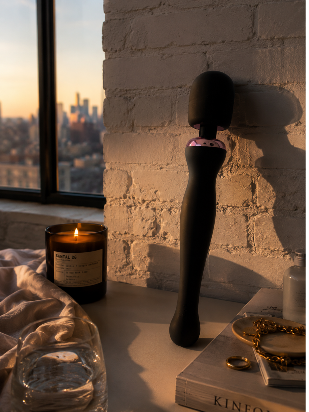 Black massage wand standing beside a candle near a window at sunset with linen and warm ambient lighting.