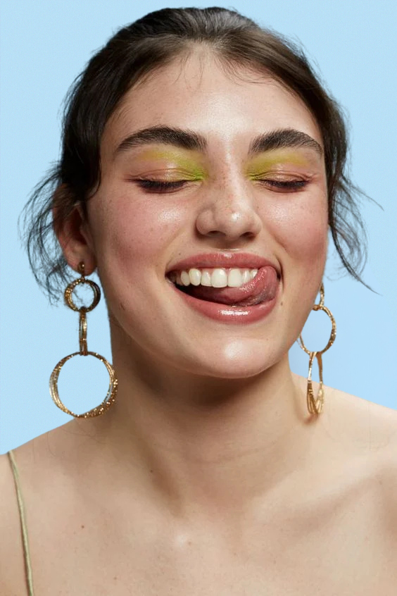 A portrait of a woman with her eyes closed, smiling playfully while sticking out her tongue and touching her upper lip. She wears bold green eyeshadow, glossy lips, and large gold hoop earrings with multiple rings. Her hair is pulled back in a loose, textured style against a light blue background.