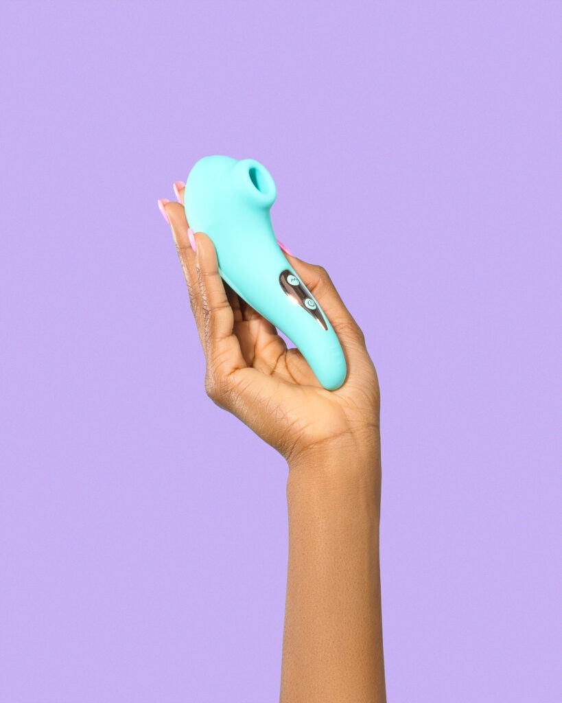 Hand holding the teal Kii pulsing-bristle clitoral stimulator against a lilac background