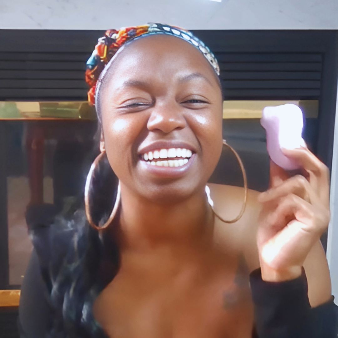Smiling intimacy educator holding a small purple clitoral stimulator toy in front of a fireplace.