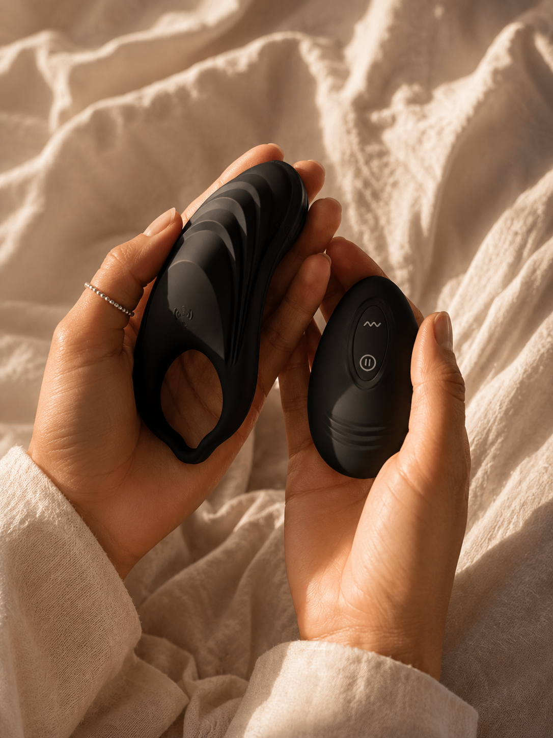 Hands holding the Esmerelda Maximus couples vibrating ring and wireless remote on soft cream bedding in warm morning light.