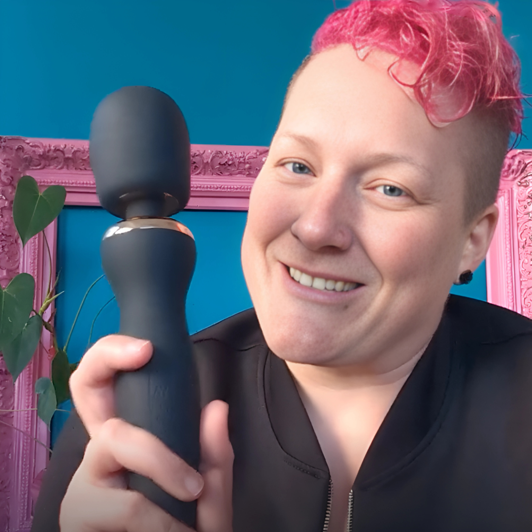 Smiling intimacy educator holding the Esmerelda Full Body Massager in front of a bright blue wall and pink frame.