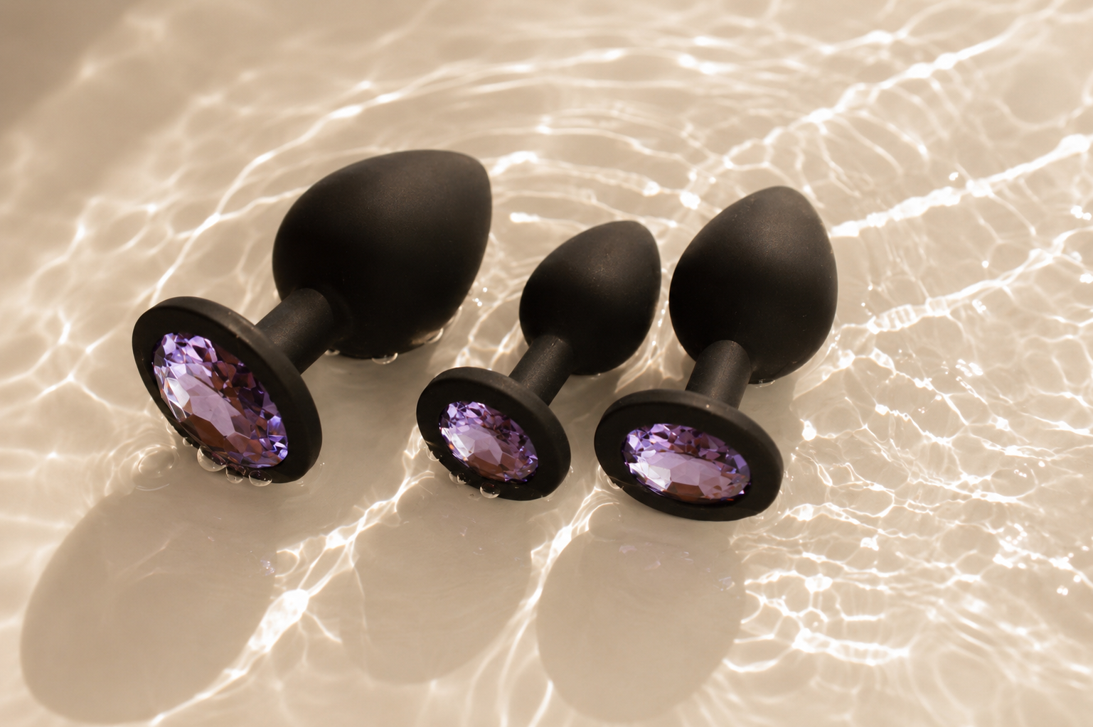 Three matte black silicone anal plugs from the Esmerelda Charlie's Plug Set rest in shallow crystal-clear water with warm sunlight creating soft ripples and reflections. Each plug features a faceted purple gemstone base and is displayed in three graduated sizes.
