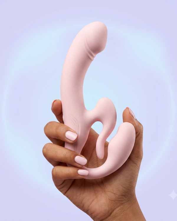 Pale pink Rosé All Day strapless couples vibrator held in a hand against a soft lavender and blue studio background.
